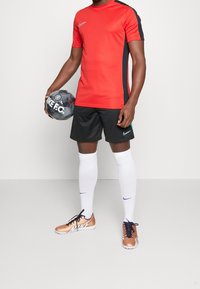 Persona vestida con ropa deportiva de Nike en rojo y negro, calcetas blancas hasta la rodilla y botas de fútbol Nike doradas, sosteniendo un balón de fútbol Nike F.C.
