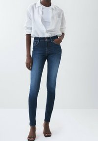 Mörkblå denim skinny jeans med hög midja, kombinerade med en vit skjorta med knappar och svarta klacksandaler, med klassisk femfickdesign.