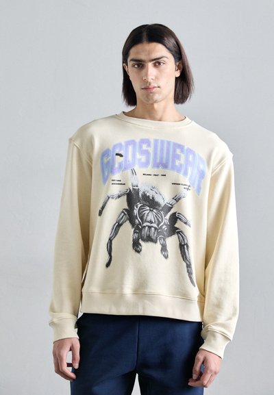 Beige sweatshirt featuring a large black spider graphic and "GCDSWEAR" in purple text. Ribbed cuffs and hem; soft, textured fabric.