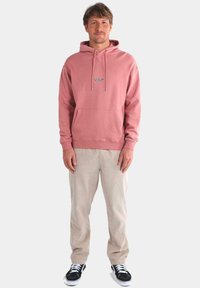 Sudadera con capucha en color rosa empolvado, con un bolsillo frontal y cierre de cordón, combinada con pantalones de pana en color crema y zapatillas negras.