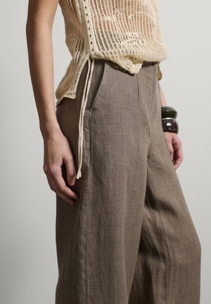 Nahaufnahme einer Person, die ein beige locker gestricktes ärmelloses Oberteil und braune Leinenhose mit weitem Bein und hoher Taille trägt, mit dunklen Armreifen am rechten Handgelenk.