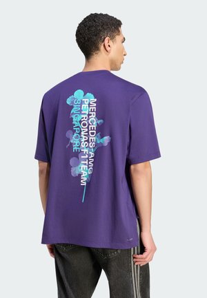 Uomo che indossa una t-shirt viola con motivo floreale blu e testo "Mercedes-AMG Petronas F1 Team Singapore" sulla schiena, abbinata a pantaloni neri.