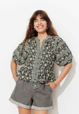 Femme souriante, portant un chemisier à motifs floraux noir et blanc avec des manches bouffantes et un short en denim gris retroussé.