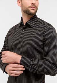 Camicia nera con sottili righe verticali dorate, design con bottoni, colletto a punta e taschino singolo. Tessuto dalla texture liscia.