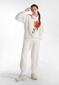 Mujer joven de pie con la mano cerca de la frente, vistiendo pantalones de chándal blancos, sudadera con estampado de flores en rojo-anaranjado y zapatillas beige con rayas verdes.