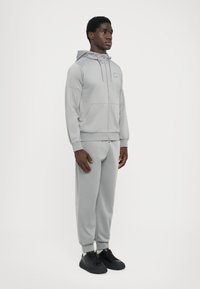 Lys grå zip-up hættetrøje med kængurulommer og matchende tapered sweatpants, lavet af blødt stof, parret med sorte sneakers.