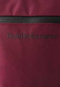 Donkerrood gestructureerde stof met een zwarte rits en het zwarte "Peak Performance" logo dat over het midden is gedrukt.