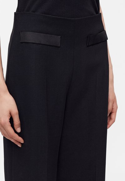 Gros plan d'une personne portant un pantalon noir sur mesure avec deux poches à rabat horizontales et un haut noir côtelé.