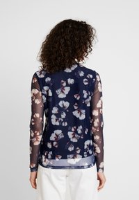 Långärmad blus i marinblått, tillverkad av genomskinligt tyg med ett blommönster i vitt och rött. Rundad nederkant, mjuk textur och figurnära silhuett.