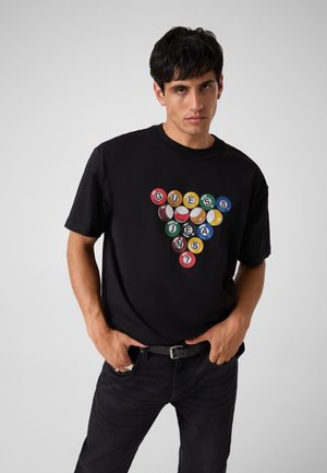 Junger Mann trägt ein schwarzes T-Shirt mit bunten Billardkugeln, die "Guess Jeans" in einer dreieckigen Formation bilden, kombiniert mit dunkler Hose und Gürtel.