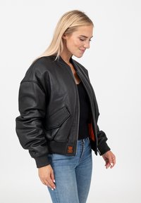 Zwarte leren bomberjack met ribgebreide manchetten en zoom. Bevat een zijzak en heeft een oranje voering. Gekleed met slim fit jeans.
