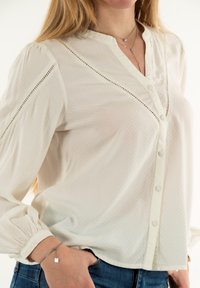 Blusa blanca de botones con escote en V, tela texturizada con un patrón de puntos y mangas fruncidas. Llevada con vaqueros azules.