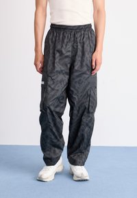 Cargo bukser i mørkegrå stof med et geometrisk mønster, udstyret med elastisk talje og flere sidelommer. Båret med hvide sneakers.