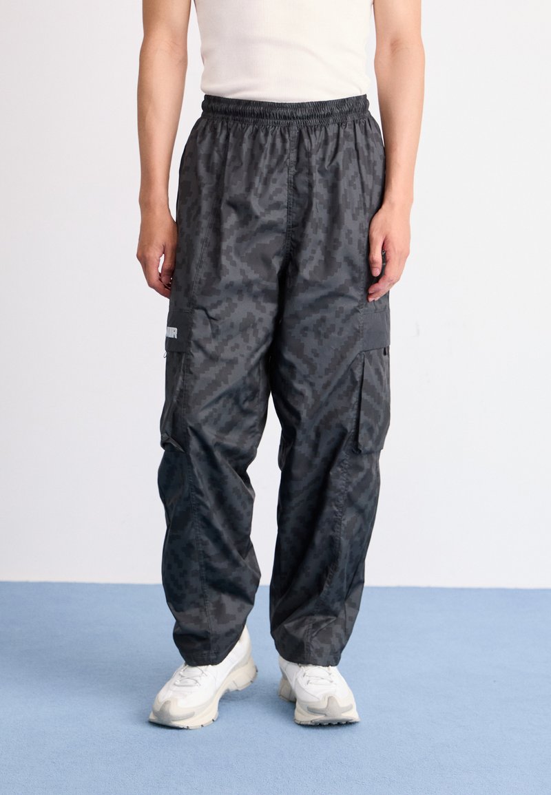 Cargo bukser i mørkegrå stof med et geometrisk mønster, udstyret med elastisk talje og flere sidelommer. Båret med hvide sneakers.