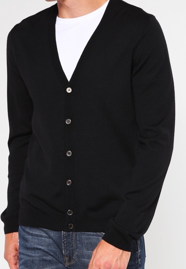 Cardigan nero a scollo a v realizzato in un morbido tessuto a maglia, caratterizzato da una fila di bottoni argentati, maniche lunghe e polsini a costine. Indossato sopra una t-shirt bianca.