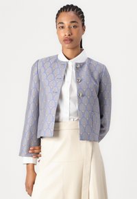 Blåmønstret cropped jakke med gyldent blomstermotiv, med rund krage og tre metallknapper foran. Hvit skjorte under.