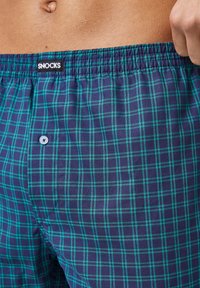 Herenboxers in marineblauw en groen geruit patroon, met een elastische tailleband en een enkele knoopdetail. Zachte stoftextuur.