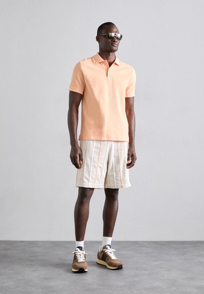 Polo shirt léger pêche, shorts rayés crème et orange, baskets brunes et lunettes de soleil. Tissu décontracté et respirant, coupe décontractée.