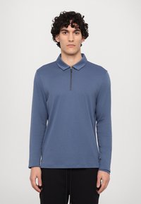 Langærmet polo-shirt i dæmpet blå, med en lynlåset krave, glat stoftekstur og afslappet pasform. Båret med mørke bukser.
