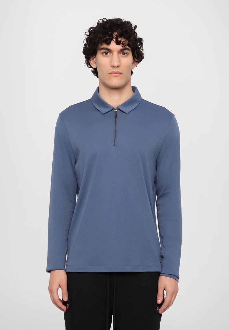 Langærmet polo-shirt i dæmpet blå, med en lynlåset krave, glat stoftekstur og afslappet pasform. Båret med mørke bukser.
