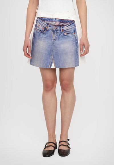 Jupe en denim avec un lavage bleu et des poches avant, superposée sur une base en tissu blanc. Présente une couture visible et une texture en denim distincte.