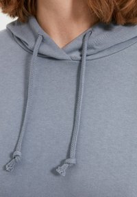 Sudadera de color gris claro con una textura en la tela, que presenta una capucha con cordón ajustable y tiras trenzadas en contraste en el escote.