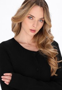 Cardigan nero con scollo rotondo, tessuto strutturato e chiusura con bottoni. Maniche lunghe che coprono parzialmente le mani, evidenziando il morbido materiale knit.