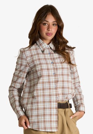 Giovane donna con lunghi capelli castani indossa una camicia a quadri marrone e bianca abbottonata, infilata in pantaloni beige con cintura scura, mano in tasca.