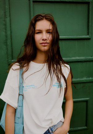 Junge Frau mit langen braunen Haaren, die ein weißes T-Shirt trägt und eine hellblaue Umhängetasche bei sich hat, steht vor einem grünen Türhintergrund.