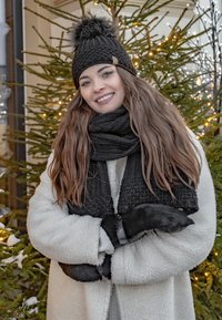 Black knitted beanie with pom-pom, matching scarf, black gloves; white textured coat. Background includes evergreen tree and soft lighting.