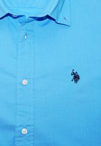 Camisa de botones azul claro, material de algodón, que presenta un logo bordado oscuro de un jinete con un martillo en el área del pecho izquierdo.