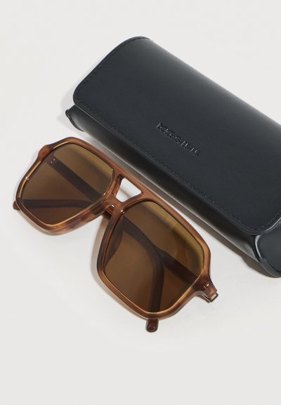 Καφέ υπερμεγέθη τετράγωνα γυαλιά ηλίου με καφέ φακούς δίπλα σε μια μαύρη σκληρή θήκη Dolce & Gabbana πάνω σε λευκή επιφάνεια.