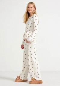 Conjunto de pijama blanco con estampado de osito de peluche marrón. Mangas largas, pantalones de corte relajado. Tejido suave, combinado con zapatillas beige peludas con detalles de orejas.