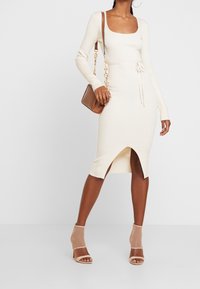 Rippstrick, langärmliges Kleid in Creme mit quadratischem Ausschnitt, Zugband in der Taille und einem Schlitz vorne. Kombiniert mit hautfarbenen Mesh-Absätzen und einer braunen Tasche.