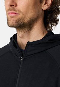 Felpa nera con zip e una consistenza morbida, con cappuccio aderente, protezione per il mento e dettagli di cucitura discreti, indossata da una persona con barba.