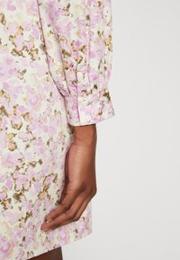 Φόρεμα με λουλουδάτο μοτίβο σε κρέμ απόχρωση, διακοσμημένο με ροζ και χρυσές λεπτομέρειες. Μακριά, συγκεντρωμένα μανίκια με κουμπί στο τελείωμα. Λείο υλικό.