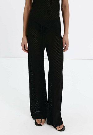 Jambes portant un pantalon en maille noire ample avec une taille à cordon et des sandales noires à bout ouvert, debout sur un sol blanc.