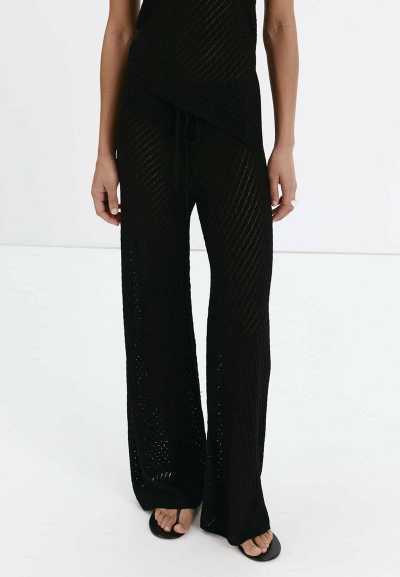 Jambes portant un pantalon en maille noire ample avec une taille à cordon et des sandales noires à bout ouvert, debout sur un sol blanc.