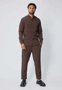 Chemise boutonnée à manches longues marron associée à un pantalon assorti. Texture lisse, coupe décontractée et chaussures plates sombres complètent le look.