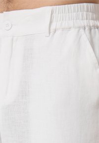 Witte linnen broek heeft een relaxte fit met een elastische tailleband en een sluiting met een knoop aan de voorkant. De stof heeft een lichte, gemêleerde weving.