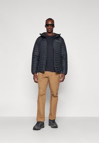 Sort pufferjakke med hætte, grå langærmet skjorte, khaki bukser og mørke sneakers; stående model iført solbriller.