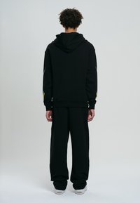 Osoba z kręconymi włosami, ubrana w czarną bluzę z kapturem i luźne czarne spodnie, stojąca plecami do obiektywu na jednolitym tle.