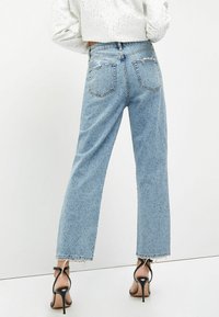Jeans de mezclilla de color azul claro con pierna recta, dobladillos deshilachados y dos bolsillos traseros, combinados con tacones altos negros con tiras.