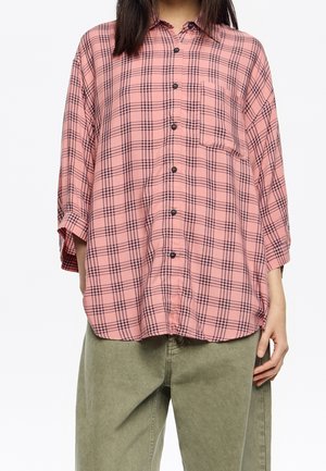 Kobieta ubrana w luźną różową koszulę w kratę z kieszenią na piersi oraz oliwkowozielone spodnie, stojąca z rozluźnionymi ramionami.