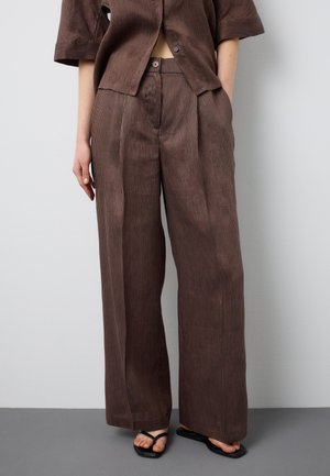 Person trägt hochtaillierte, braune Nadelstreifen-Hosen mit weitem Bein, dazu ein passendes Hemd mit Knöpfen und schwarze Sandalen mit quadratischer Zehenpartie.