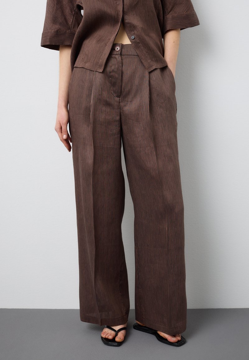 Person trägt hochtaillierte, braune Nadelstreifen-Hosen mit weitem Bein, dazu ein passendes Hemd mit Knöpfen und schwarze Sandalen mit quadratischer Zehenpartie.