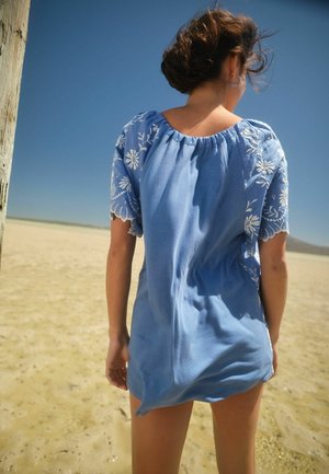 Donna con capelli corti che indossa un vestito blu con ricami floreali bianchi, in piedi su una spiaggia soleggiata, girata di spalle rispetto alla fotocamera.