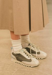 Sneaker con una toma bianca e grigia caratterizzata da una finitura testurizzata, dettagli blu chiaro e suole bianche seghettate. Indossate con calze bianche.
