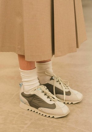 Sneaker con una toma bianca e grigia caratterizzata da una finitura testurizzata, dettagli blu chiaro e suole bianche seghettate. Indossate con calze bianche.