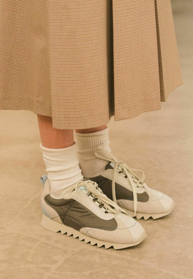 Sneaker con una toma bianca e grigia caratterizzata da una finitura testurizzata, dettagli blu chiaro e suole bianche seghettate. Indossate con calze bianche.